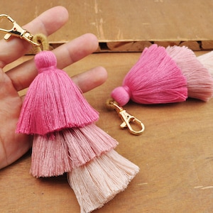 Könnte beinhalten: Zwei rosa und weiße Quasten-Schlüsselanhänger mit goldenen Metallschließen. Die Quasten sind übereinander gelegt und haben einen Farbverlaufseffekt.