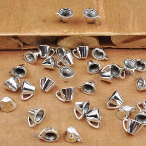 Puede incluir: Una colección de abalorios de taza de té de metal plateado. Los abalorios son pequeños y detallados, con un diseño realista de taza de té.