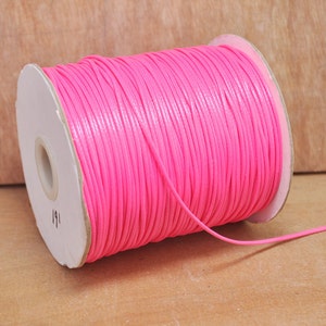 Pode incluir: Uma bobina de cordão de algodão encerado de cor rosa brilhante. O cordão está enrolado em torno de uma bobina de cartão branco com uma etiqueta que diz "11".