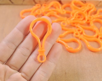 May include: A close-up of a hand holding a bright orange plastic key chain clip. The clip has a small loop at the top and a larger loop at the bottom. There are many more of these clips in the background.