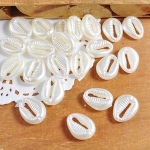 May include: A collection of white, smooth, shell-shaped beads with a hole in the centre. The beads are scattered on a white lace doily and a wooden surface.