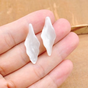 Puede incluir: Dos abalorios de plástico de forma de hoja, de color blanco nacarado. Los abalorios miden aproximadamente 5 cm de largo y tienen una superficie ligeramente texturizada.