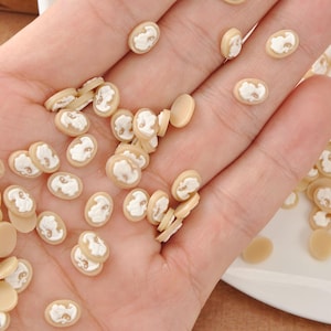 May include: A collection of small, oval-shaped cameo beads in a light beige color. Each bead features a white relief portrait. The beads are scattered across a hand and a white plate, showcasing their delicate design.