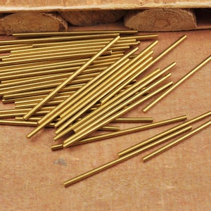 May include: A pile of gold-colored metal rods, likely brass, arranged on a brown wooden surface. The rods are all the same length and diameter, and they are stacked in a random pattern.
