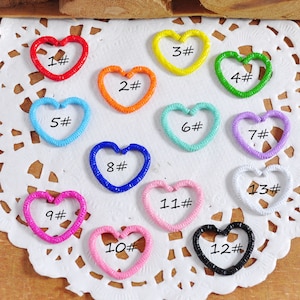 May include: Thirteen heart-shaped metal charms in various colors, numbered 1 through 13. The charms are arranged in a circle on a white lace background.