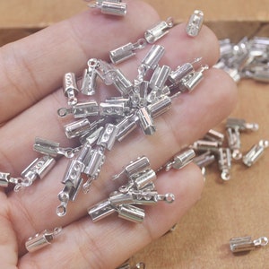 May include: A close-up of a hand holding a large quantity of silver metal crimp end caps. The caps are used for jewelry making and have a small loop on one end.