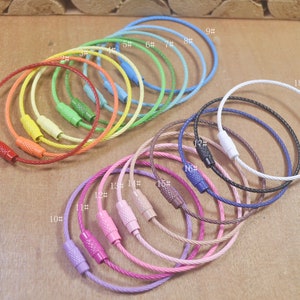 Könnte beinhalten: Sortiment von 18 bunten Schlüsselringen aus Metallkabel. Die Schlüsselringe sind in einem Kreis angeordnet, wobei die Zahlen 1 bis 18 neben jedem Ring gedruckt sind. Die Schlüsselringe sind in verschiedenen Farben erhältlich, darunter Rot, Orange, Gelb, Grün, Blau, Lila und Weiß.