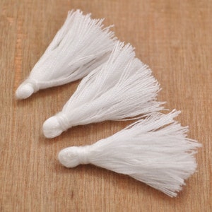 May include: Three white cotton tassels, each with a long, fluffy fringe. The tassels are arranged in a row on a wooden surface.