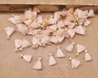 May include: A large group of small, light pink tassels with gold accents. The tassels are scattered on a brown surface.