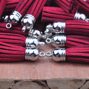 May include: A group of burgundy suede tassels with silver metal caps and loops. The tassels are arranged on a wooden surface.