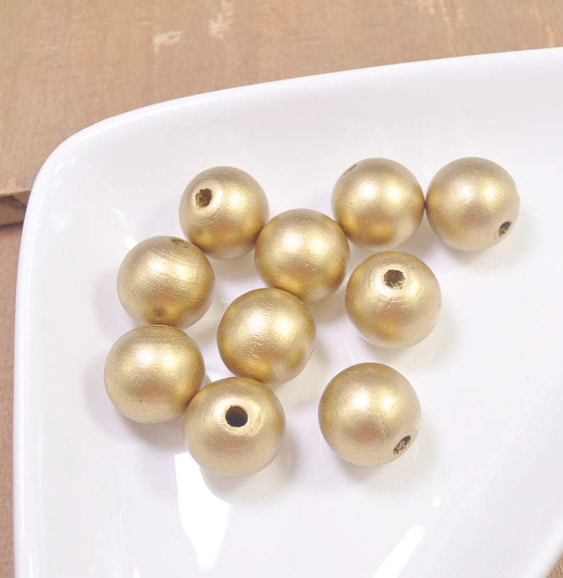 Peut inclure: Dix perles en bois dor&eacute;es. Les perles sont rondes et ont une finition lisse et brillante. Elles sont dispos&eacute;es de mani&egrave;re al&eacute;atoire sur une surface blanche.