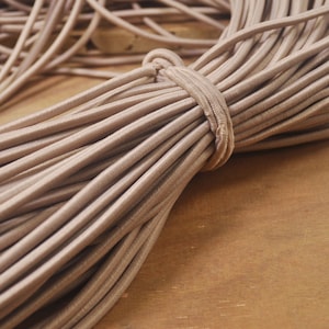 Puede incluir: Primer plano de un paquete de cordón elástico beige. El cordón es suave y flexible, ideal para proyectos de manualidades.