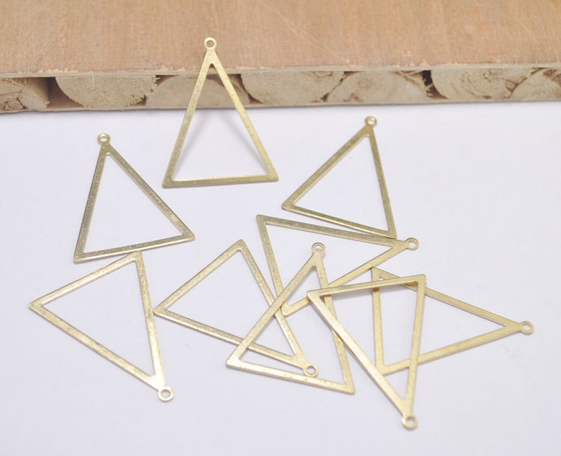 May include: A collection of brass-colored triangle charms. Each charm has a triangular shape with a small loop at the top and a hole at the bottom for attaching to jewelry. The charms are scattered on a white surface.