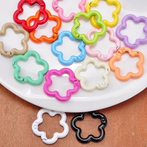 May include: A collection of flower-shaped spring gate rings in various colors including red, orange, yellow, green, blue, purple, pink, black, white, and beige. Each metal ring features a hinged opening mechanism for attaching charms or keys.