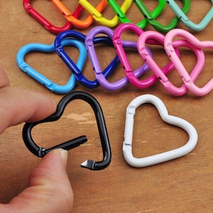 May include: A collection of heart-shaped carabiner clips in various colors, including red, orange, yellow, green, blue, purple, pink, black, and white. One black clip is held by a hand. These decorative clips are suitable for keychains or other craft projects.