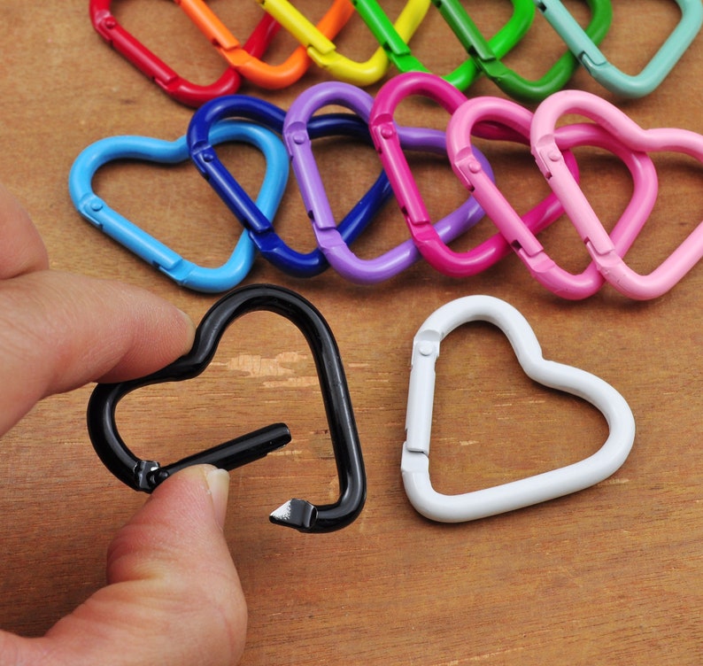May include: A collection of heart-shaped carabiner clips in various colors, including red, orange, yellow, green, blue, purple, pink, black, and white. One black clip is held by a hand. These decorative clips are suitable for keychains or other craft projects.