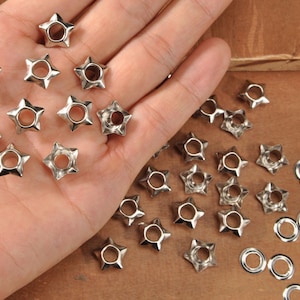 May include: Silver-colored star-shaped metal rivets with a central hole, scattered on a wooden surface. Several rivets are held in a hand, alongside loose rivets and silver eyelets. These rivets are ideal for crafting and adding decorative elements.