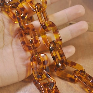 Puede incluir: Primer plano de una cadena hecha de plástico translúcido con estampado de carey. Los eslabones de la cadena son rectangulares y están conectados, creando un accesorio decorativo y audaz. Los cálidos tonos marrones y ámbar le dan un aspecto vintage.