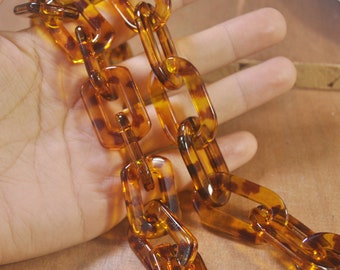 May include: A close-up of a chain made of translucent, tortoiseshell-patterned plastic. The chain links are rectangular and connected, creating a bold, decorative accessory. The warm brown and amber tones give it a vintage look.