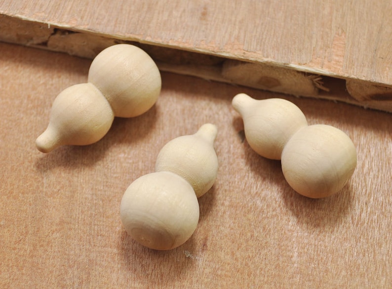 May include: Three wooden gourds, each with two rounded sections, are arranged on a light brown wooden surface. The gourds are unpainted and have a natural wood finish.