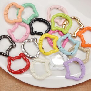 Puede incluir: Una colecci&oacute;n de ganchos de cierre met&aacute;licos con forma de gato en varios colores, incluyendo rosa, verde, negro y naranja. Los cierres est&aacute;n dispuestos sobre un plato blanco, mostrando su acabado brillante y dise&ntilde;o &uacute;nico. Ideales para manualidades y bisuter&iacute;a.