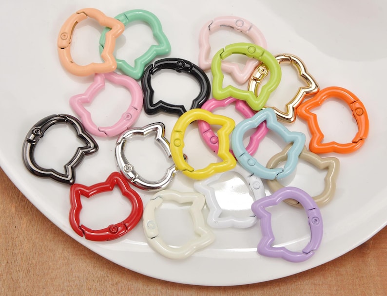 Puede incluir: Una colecci&oacute;n de ganchos de cierre met&aacute;licos con forma de gato en varios colores, incluyendo rosa, verde, negro y naranja. Los cierres est&aacute;n dispuestos sobre un plato blanco, mostrando su acabado brillante y dise&ntilde;o &uacute;nico. Ideales para manualidades y bisuter&iacute;a.