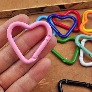 May include: A collection of heart-shaped carabiner clips in various colors, including pink, blue, red, green, orange, and black. The clips are made of metal and have a spring-loaded gate for secure attachment. The background is a wooden surface.