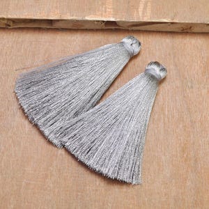 May include: Two silver-gray tassels with a silky texture are displayed against a light brown wooden surface. The tassels are tied at the top with a matching silver thread, and the strands fan out to create a full, decorative shape. These tassels are suitable for various craft projects.