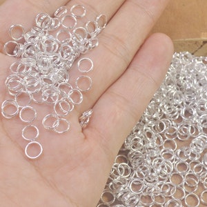 Könnte beinhalten: Ein großer Haufen silberner Sprungringe, eine Art Schmuckzubehör, wird in einer Hand gezeigt. Die Ringe sind klein und rund und perfekt zum Schmuckmachen.