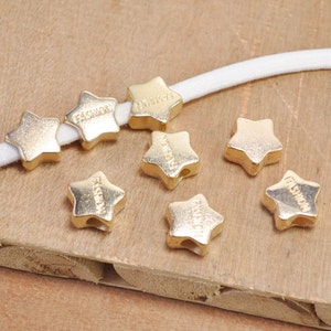 May include: Gold star-shaped beads with the word "FASHION" engraved on them. The beads are strung on a white elastic cord.