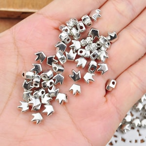 May include: A collection of small, silver-colored metal crown-shaped beads. The beads are scattered across a surface, with a few held in a hand. Each bead has a crown design with three points and a hole for stringing.