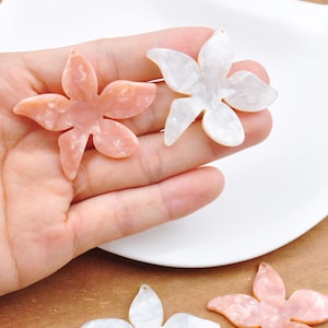 Peut inclure: Deux breloques en forme de fleur en plastique, l'une blanche et l'autre rose, avec un effet marbré. Les breloques sont en forme d'étoile à cinq branches avec un trou au centre pour les attacher à des bijoux.