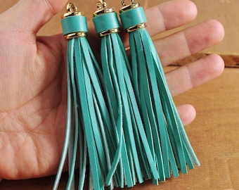 May include: Three teal leather tassels with gold metal tops. The tassels are hanging from a hand.