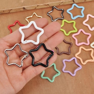 Pode incluir: Uma variedade de fechos metálicos em forma de estrela em várias cores, incluindo vermelho, branco, preto, prata, dourado, verde, azul, rosa e castanho. Os fechos estão abertos e prontos para serem usados em chaveiros ou projetos de artesanato.