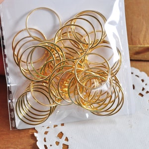 May include: A close-up of a package of gold jump rings. The rings are arranged in a circular pattern and are all the same size. The rings are in a clear plastic bag.
