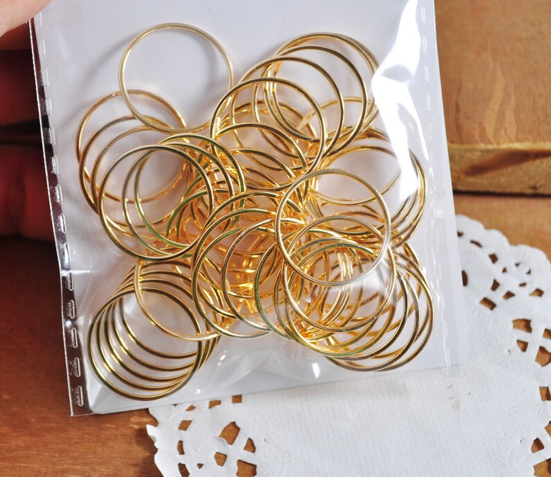 May include: A close-up of a package of gold jump rings. The rings are arranged in a circular pattern and are all the same size. The rings are in a clear plastic bag.
