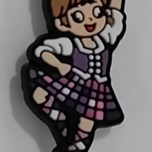May include: A cartoon charm depicting a dancer in a Scottish kilt. The figure has brown hair and is wearing a purple and white outfit with a plaid skirt. The charm is black with white and pink accents.