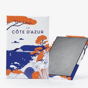 Peut inclure: Un aimant rectangulaire avec une scène côtière et le texte "LA CÔTE D'AZUR". Le design comprend des arbres orange, de l'eau bleue et un voilier blanc. L'aimant est partiellement ouvert, révélant une surface grise.