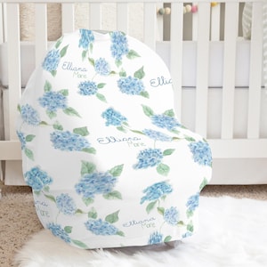 Puede incluir: Una funda blanca para asiento de coche de bebé con un estampado floral de hortensias azules. La funda tiene el nombre "Elliana Mare" en cursiva. La funda está sobre una cuna blanca y una alfombra blanca y esponjosa.
