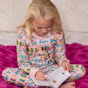 May include: A young person wearing a pink pajama set with a floral and bookshelf pattern, reading a book. The pajamas have the name "Dayton" on the chest. The book is open, and the person is pointing at the page. The background is a pink blanket.