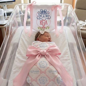 A newborn baby girl is peacefully sleeping in a hospital bed, wrapped in a pink blanket with a large pink ribbon.