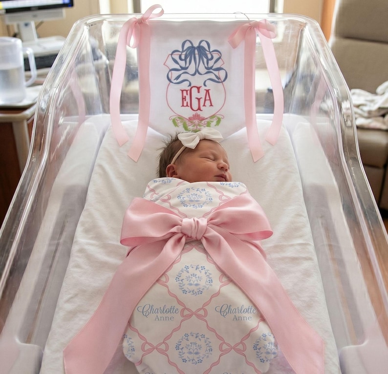 A newborn baby girl is peacefully sleeping in a hospital bed, wrapped in a pink blanket with a large pink ribbon.