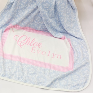 May include: A light blue baby blanket with a white floral pattern. The blanket has a pink and white gingham border with the names "Chloe Evelyn" embroidered in pink.