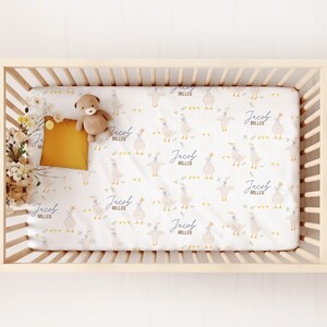 May include: A crib with a fitted sheet featuring a pattern of geese and the name "Jacob Miller" in blue script. A small teddy bear and a mustard-colored pillow rest on the sheet. The crib is made of light-colored wood.