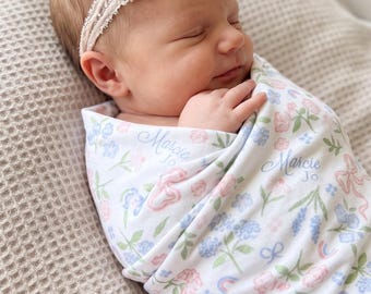May include: A newborn baby wrapped in a floral printed swaddle blanket, with the name "Marcie Jo" printed on it. The baby wears a headband with pink, blue, and cream-colored flowers. The baby is sleeping on a beige waffle-knit blanket.