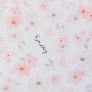May include: White fabric with a delicate floral pattern in shades of pink and gray. The word "Emery" is written in cursive script. The design features watercolor-style flowers.