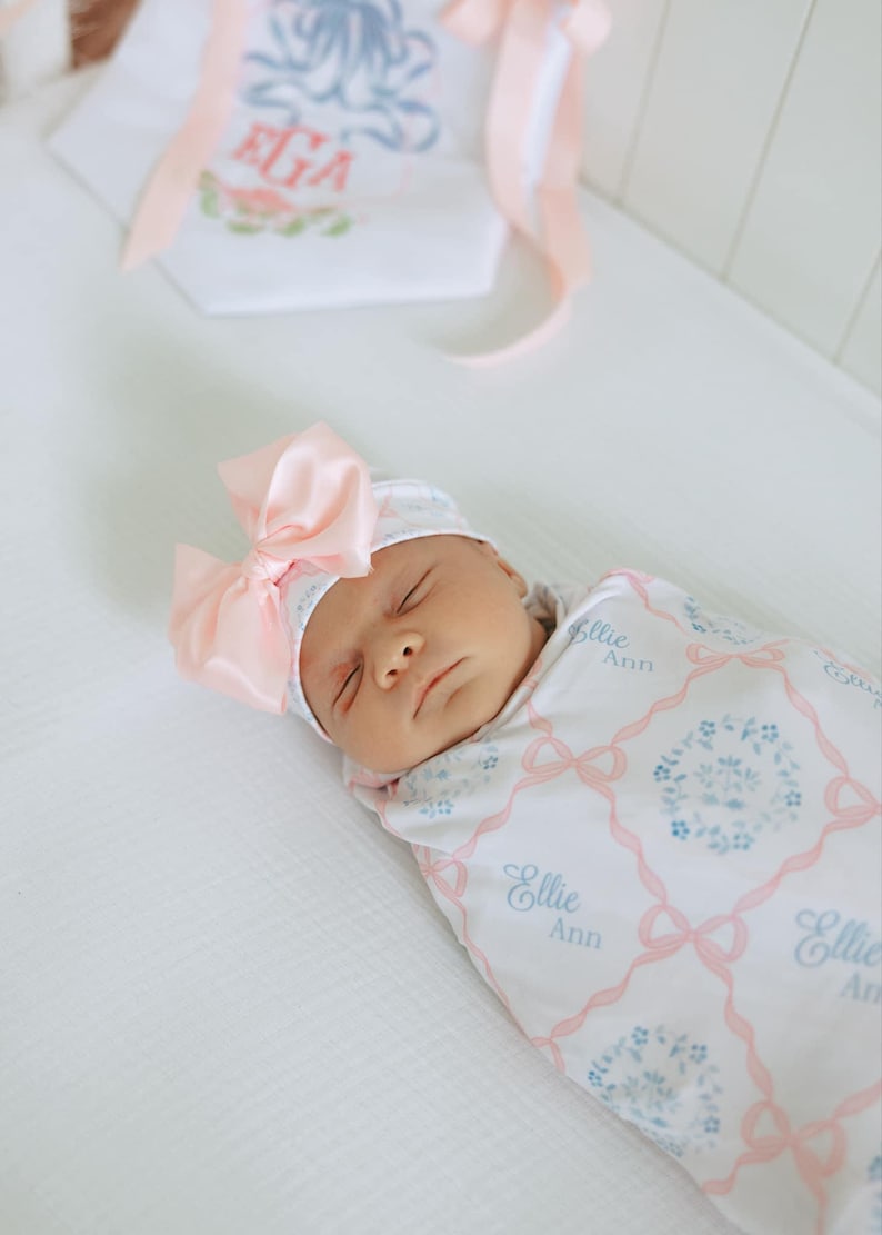 Pode incluir: Um rec&eacute;m-nascido envolto em um cobertor branco e usando um chap&eacute;u combinando, ambos adornados com um la&ccedil;o rosa e um padr&atilde;o floral. O cobertor e o chap&eacute;u apresentam o nome "Ellie Ann" em uma escrita azul suave.