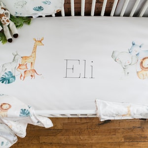 May include: White crib sheet with a watercolor design of a giraffe, zebra, lion, rhino, and elephant. The sheet also has the name "Eli" written in black.