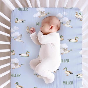 May include: A light blue baby crib sheet with a white and green duck pattern. The sheet has the name "MILLER" printed on it multiple times. A baby is sleeping on the sheet.