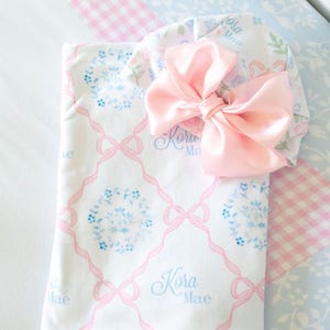 A pink and white baby blanket with a large pink bow and the name "Kora Mae" printed on it, placed on a blue and white checkered bedspread.